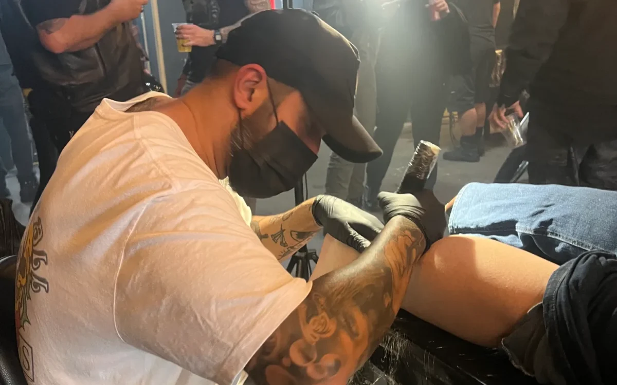 Homem com tatuagens em camiseta clara e boné preto realiza procedimento em pessoa sentada, cercado por espectadores em ambiente interno