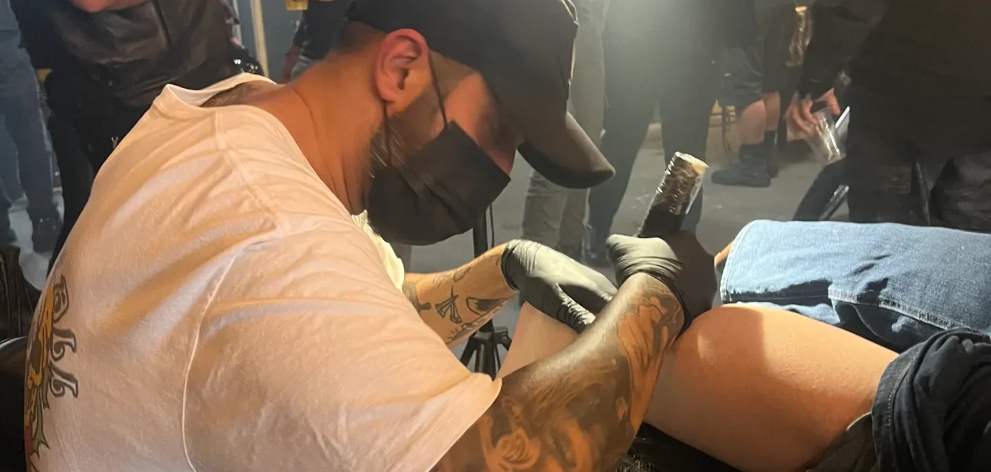 Homem com tatuagens em camiseta clara e boné preto realiza procedimento em pessoa sentada, cercado por espectadores em ambiente interno