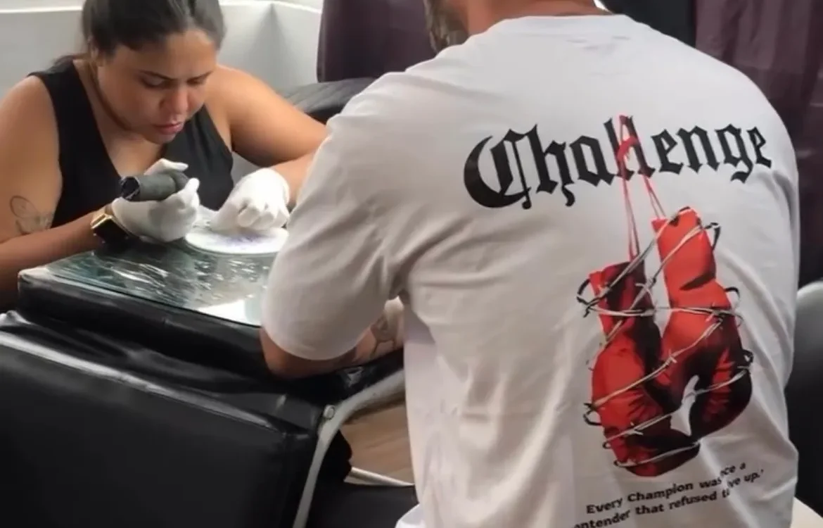 Duas pessoas em um interior, uma sentada aplicando algo na mão da outra que está de costas usando camiseta branca com texto e imagem vermelha