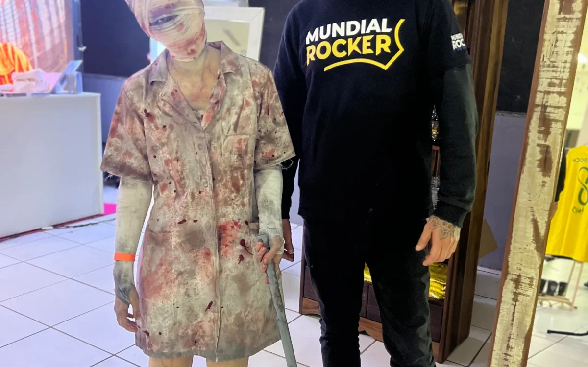 Homem com boné e moletom preto ao lado de uma figura de cera coberta com tecido translúcido, segurando uma bengala, em ambiente interno