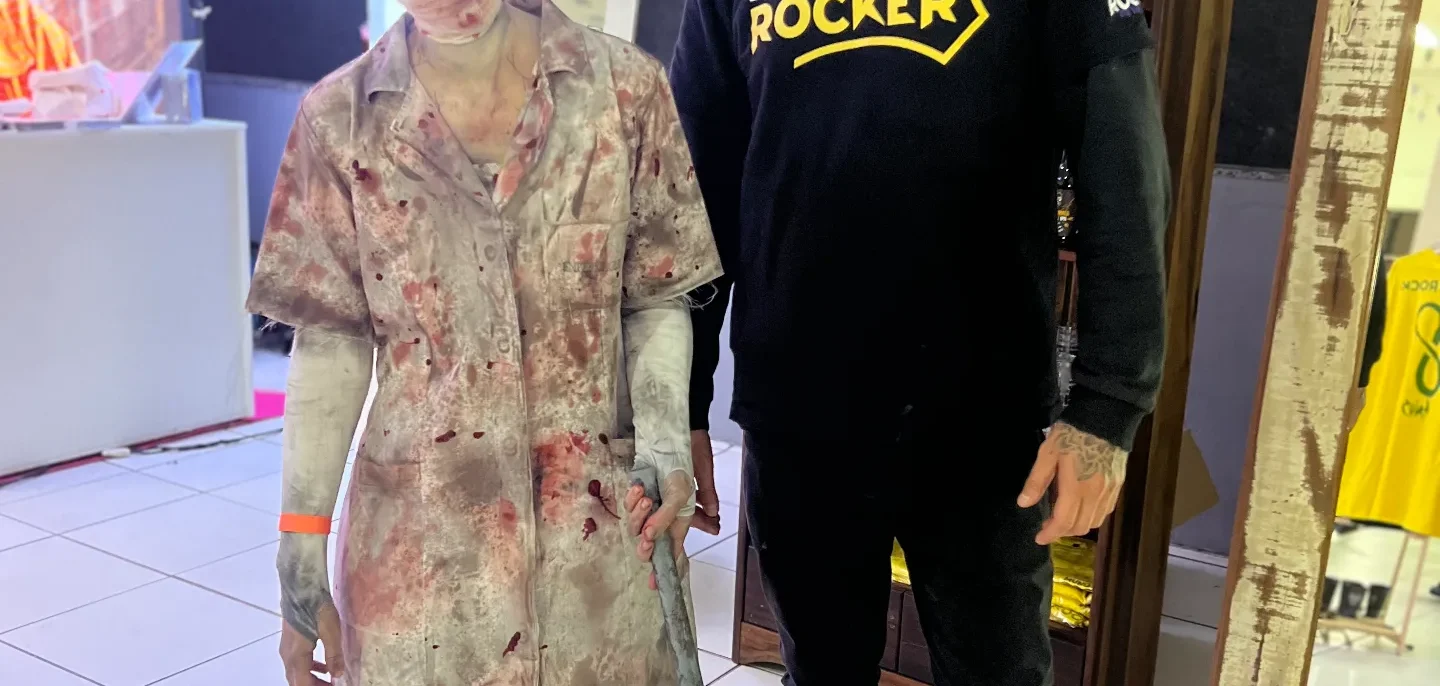 Homem com boné e moletom preto ao lado de uma figura de cera coberta com tecido translúcido, segurando uma bengala, em ambiente interno