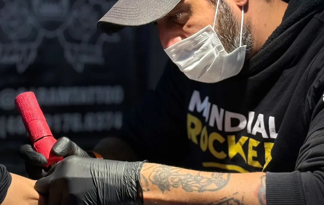 Homem com boné cinzento, máscara branca e luvas pretas, segurando um objeto vermelho sobre uma superfície escura, com tatuagem visível no braço