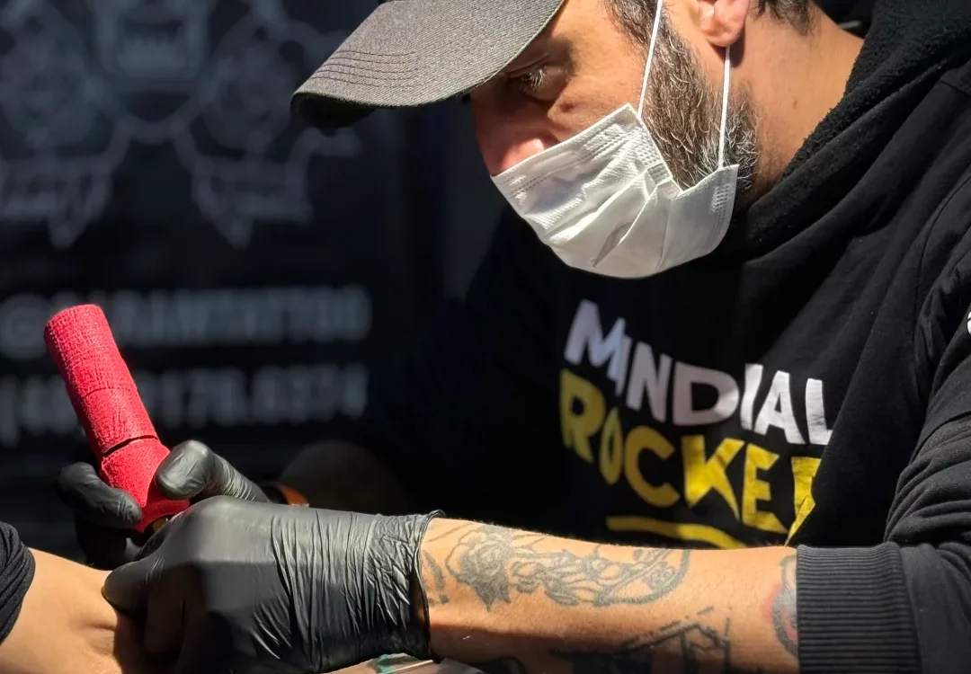 Homem com boné cinzento, máscara branca e luvas pretas, segurando um objeto vermelho sobre uma superfície escura, com tatuagem visível no braço