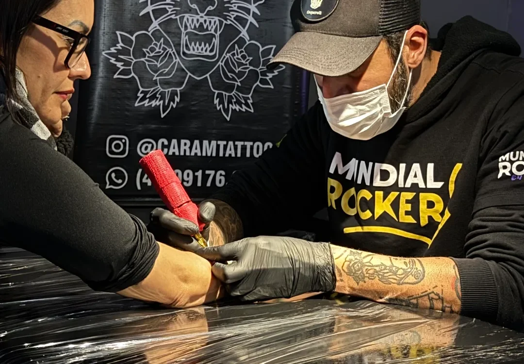 Duas pessoas em um estúdio com fundo cinzento, uma delas tatuando o braço da outra sobre uma maca coberta de plástico preto, com banner de tigre ao fundo