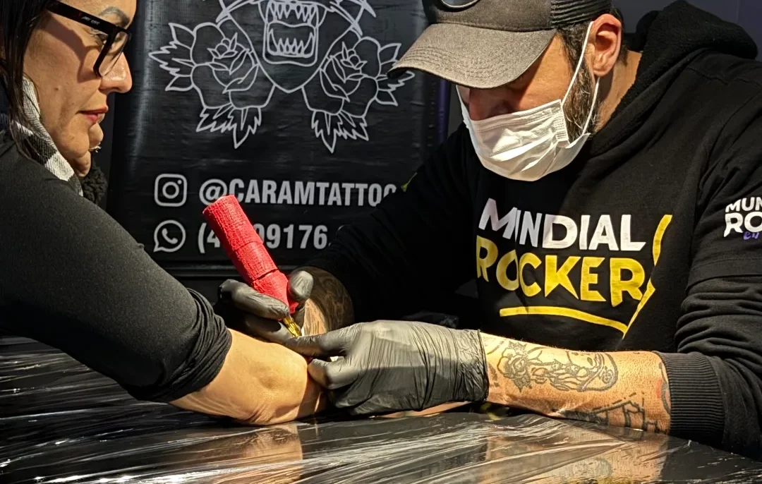 Duas pessoas em um estúdio com fundo cinzento, uma delas tatuando o braço da outra sobre uma maca coberta de plástico preto, com banner de tigre ao fundo