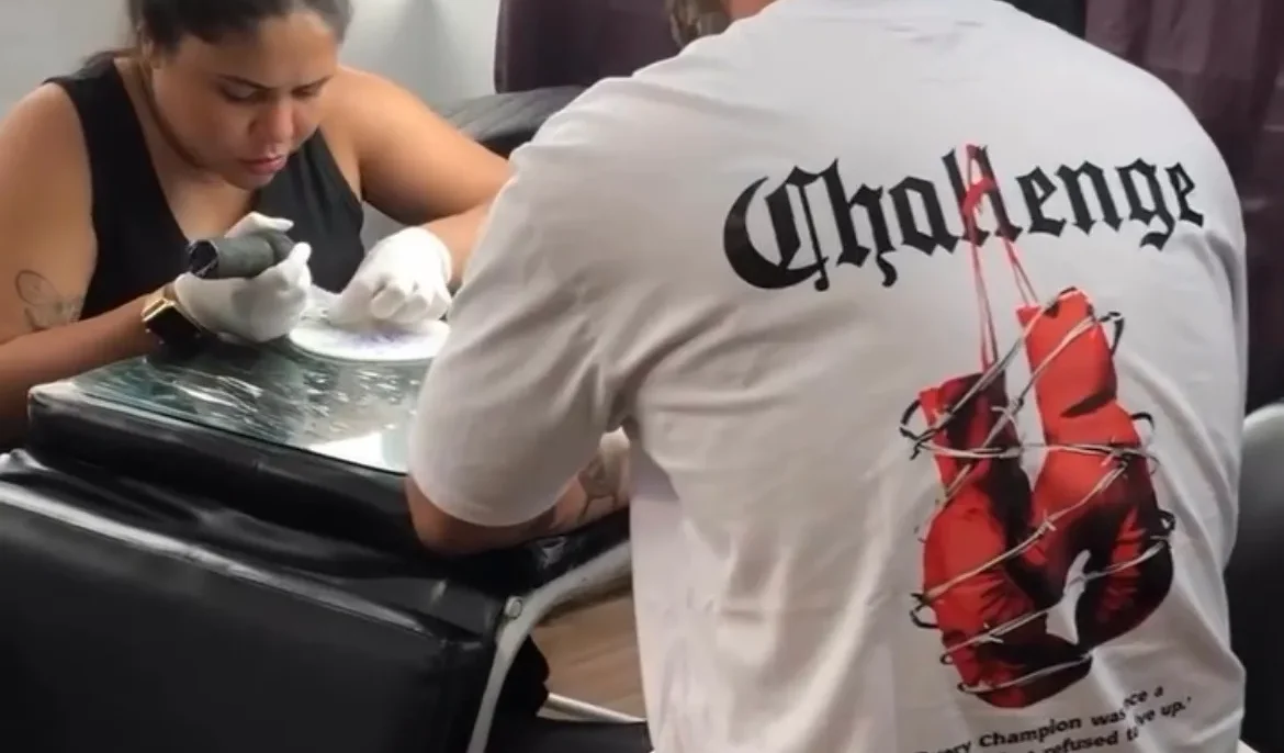 Duas pessoas em um interior, uma sentada aplicando algo na mão da outra que está de costas usando camiseta branca com texto e imagem vermelha