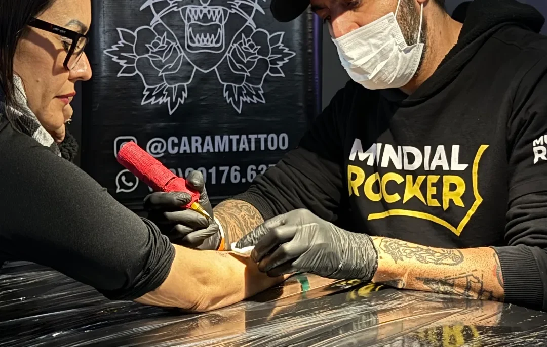 Tatuador com máscara e luvas pretas trabalha no braço de uma mulher com óculos em frente a banner com logo de leão e texto "tattoo piercing