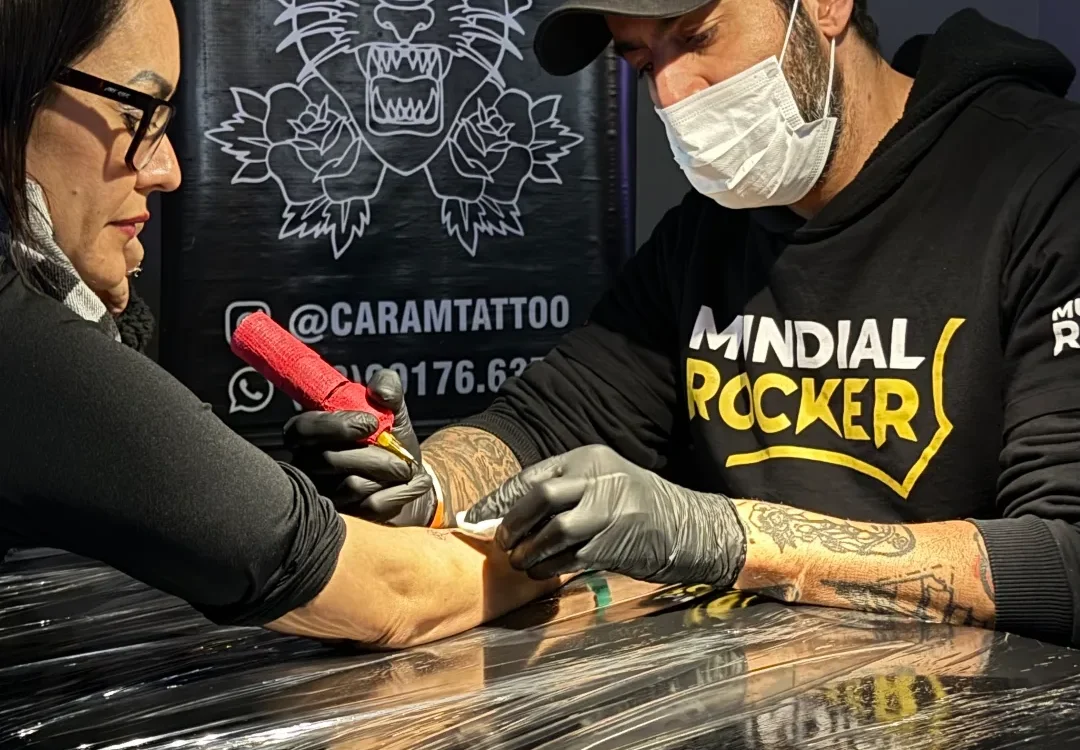 Tatuador com máscara e luvas pretas trabalha no braço de uma mulher com óculos em frente a banner com logo de leão e texto "tattoo piercing