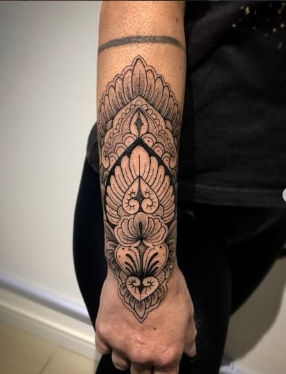Tatuagem em preto e cinza no braço com padrões geométricos e florais simétricos, detalhando mandala e ornamentos decorativos