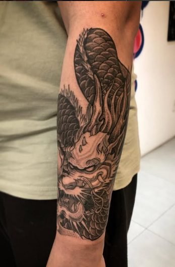 Tatuagem em preto e cinza no antebraço de uma pessoa, representando uma criatura mitológica com asas, chifres e detalhes ornamentais complexos