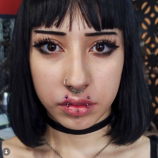 Mulher com cabelo preto liso e franja, olhos castanhos com cílios alongados, sobrancelha tatuada, piercings no nariz e boca, usando choker preto