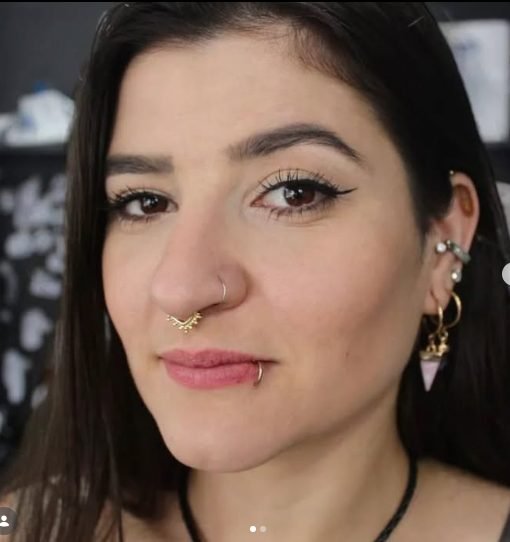 Mulher com cabelo preto longo, maquiagem com delineador, piercing no nariz e piercings nas orelhas, olhando para o lado