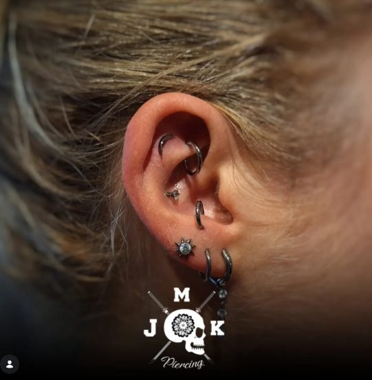 Orelha com múltiplos piercings em diferentes posições, incluindo argola no lóbulo e piercing industrial, com logotipo de estúdio de piercing ao fundo