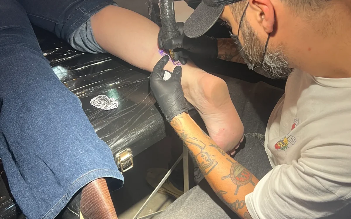 Tatuador com luva preta usando máquina de tatuar no braço de uma pessoa sentada em ambiente fechado com bebidas ao fundo