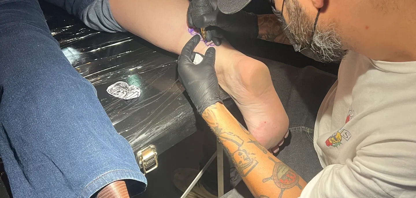 Tatuador com luva preta usando máquina de tatuar no braço de uma pessoa sentada em ambiente fechado com bebidas ao fundo