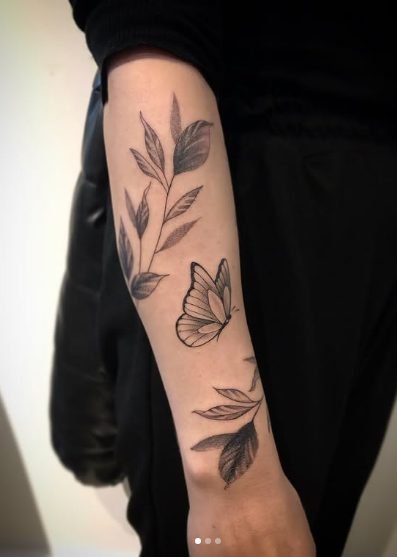 Tatuagem no braço com desenhos de plantas, folhas e uma borboleta em estilo minimalista preto e cinza